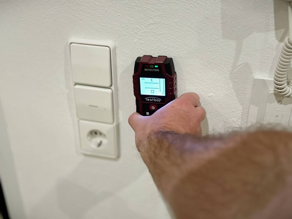 Eine Hand hält den Leitungssucher von Testboy an eine Wand neben einen Lichtschalter.