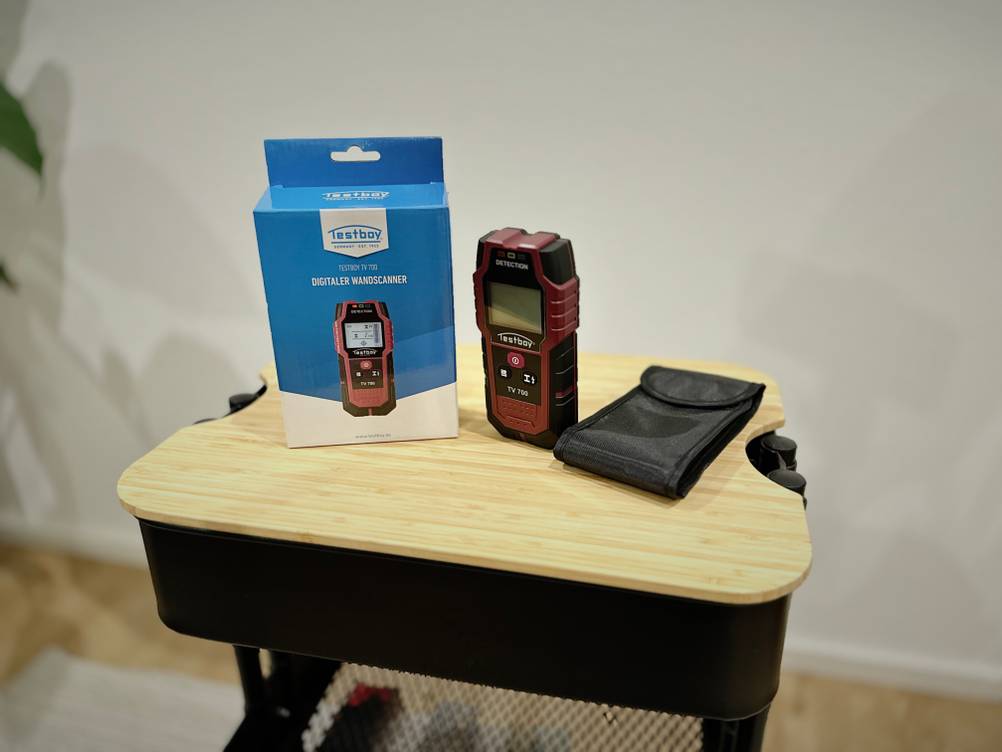 Das Produkt von Testboy steht mit der Verpackung und Zubehör auf einem hölzernen Untergrund vor einer weißen Wand.