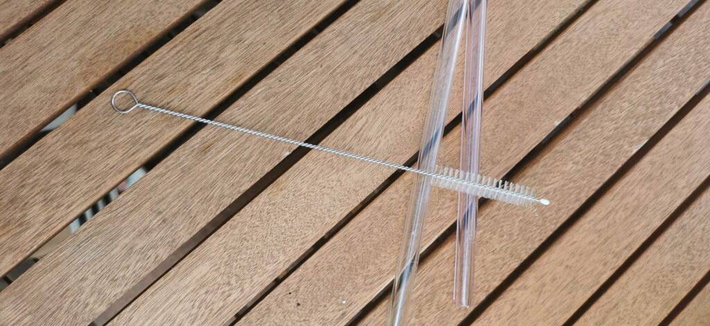 Eine kleine Reinigungsbürste liegt auf zwei transparenten Glas-Strohalmen im Test, die sich auf einer Holzoberfläche befinden.