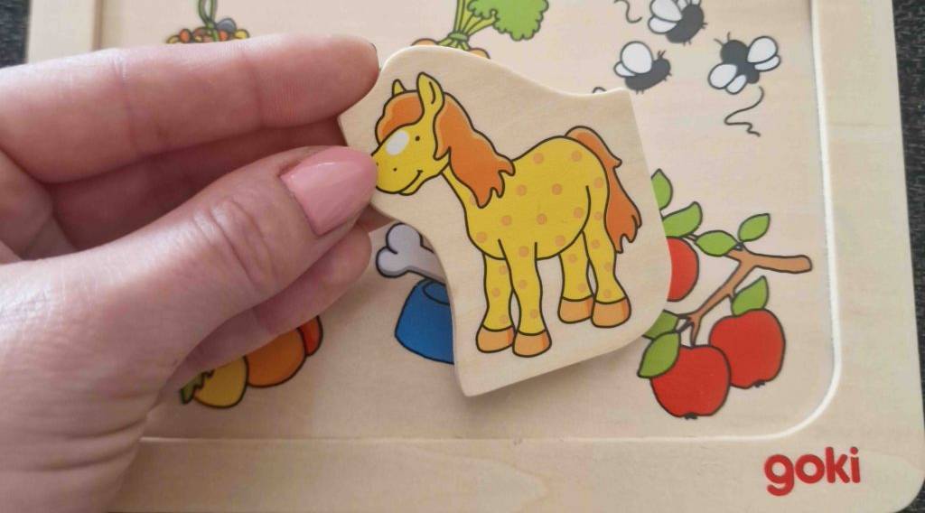 Holz-Lernspielzeug-Test: Hand hält Goki Puzzleteil mit bunter Tierzeichnung über vorgestanztem Holztablett.