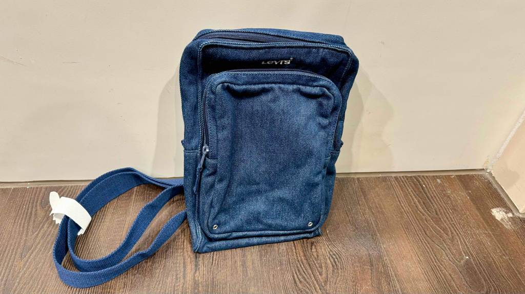 Sling-Bag-Test: ein blaues Jeans-Modell lehnt an einer beigefarbenen Wand.