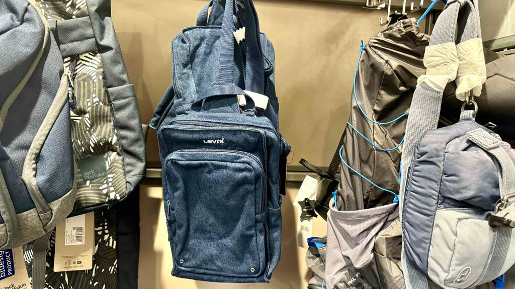 Sling-Bags im Test: ein blaues Jeans-Modell hängt zwischen mehreren Rucksäcken an einem Verkaufsstand.
