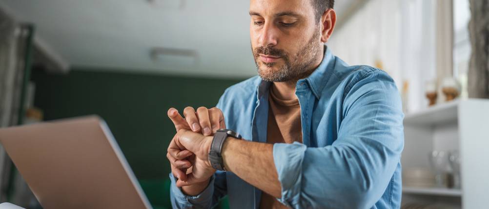 LIGE-Smartwatches im Test: junger Mann, der vor seinem aufgeklappten Laptop sitzt und auf seine Smartwatch schaut