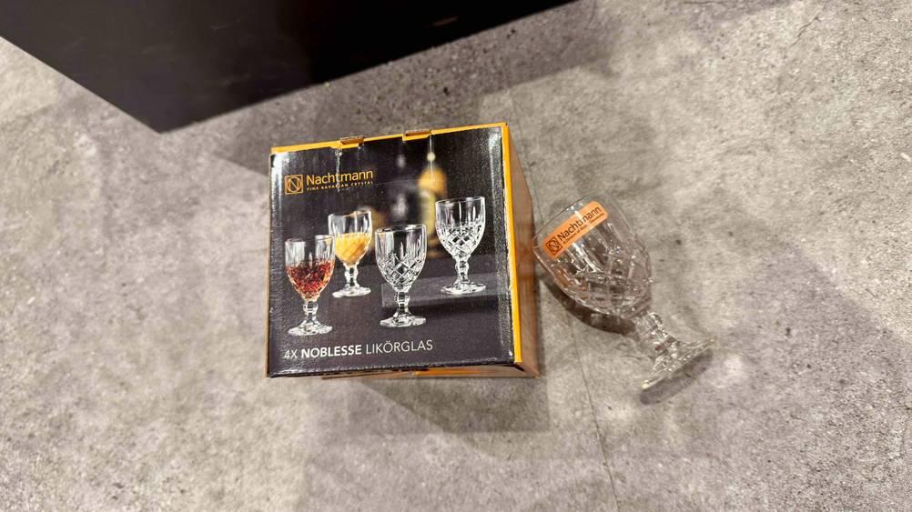 Neben der Originalverpackung der getesteten Likörgläser liegt ein einzelnes Glas auf dem Boden.