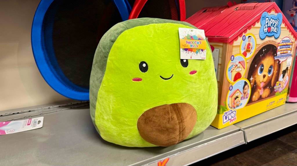 Ein gelbes Squishmallow mit Gesicht steht zum testen neben einem anderen Spielzeug in einem Regal.