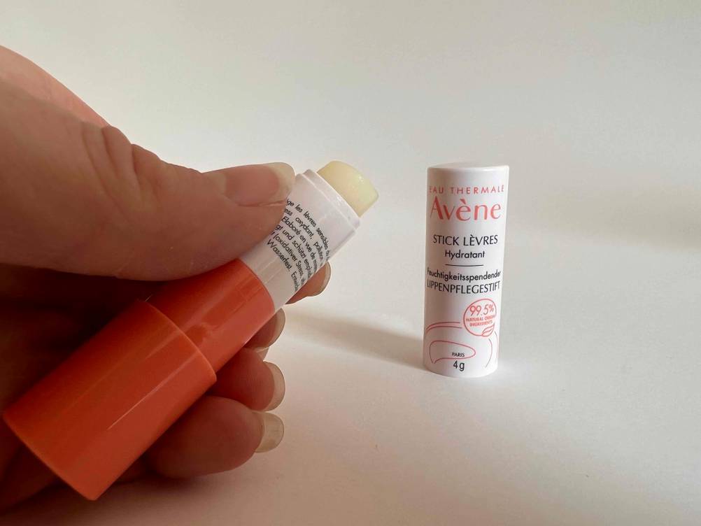Eine Hand hält eine Lippenpflege im Test neben den Deckel auf einer hellen Fläche.