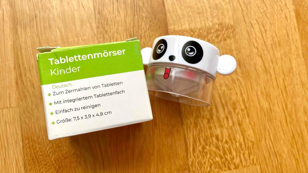 Ein getesteter Tablettenmörser im Mickey Mouse-Design liegt neben einer grün-weißen Verpackung, inklusive Produktangaben, auf einer Holzfläche.