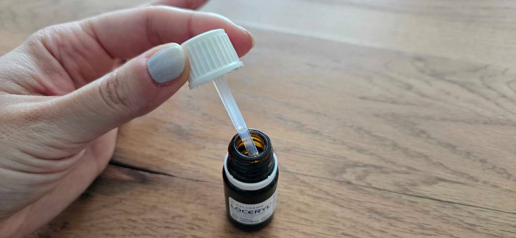 Loceryl-Mittel gegen Nagelpilz im Test: Hand hält weißen Drehverschluss mit integrierter Pipette über einer kleinen, lichtdichten Braunglas-Flasche auf Holzhintergrund.