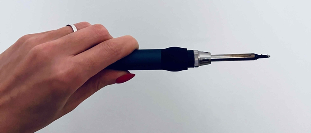 Eine Hand hält einen getesteten Lötkolben horizontal vor einer hellen Wand.