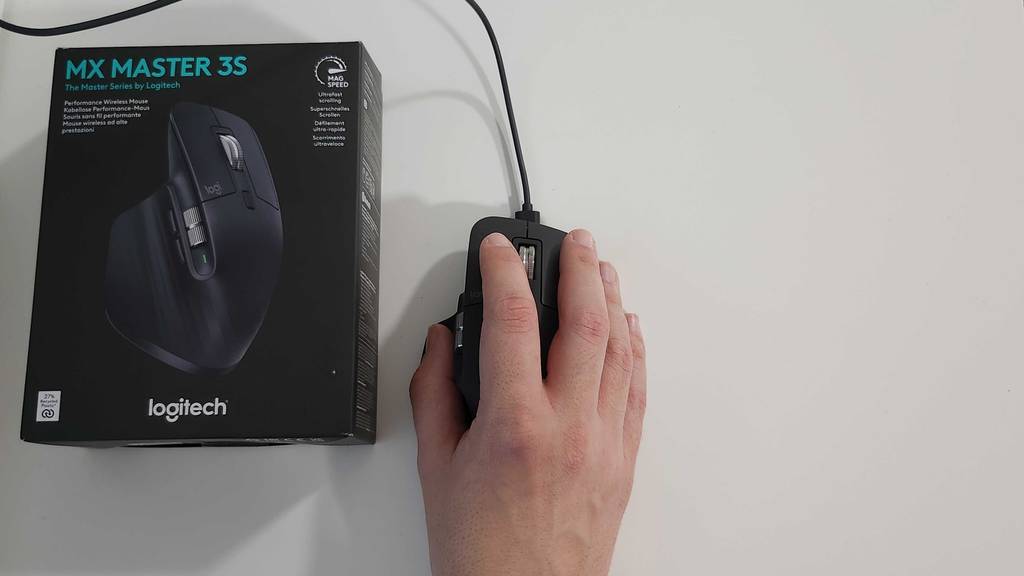 Eine Herrenhand, die eine getestete ergonomische Maus, inklusive Kabel neben einer Produktverpackung, von oben berührt.