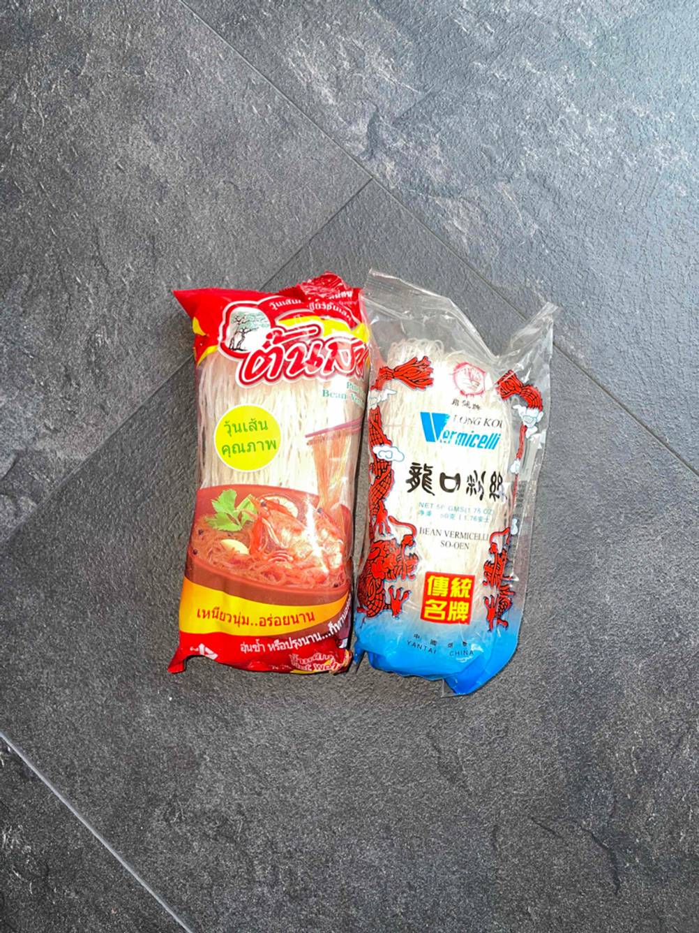 Zwei verschiedene Packungen Glasnudeln liegen zum Testen auf schwarzer Fläche.