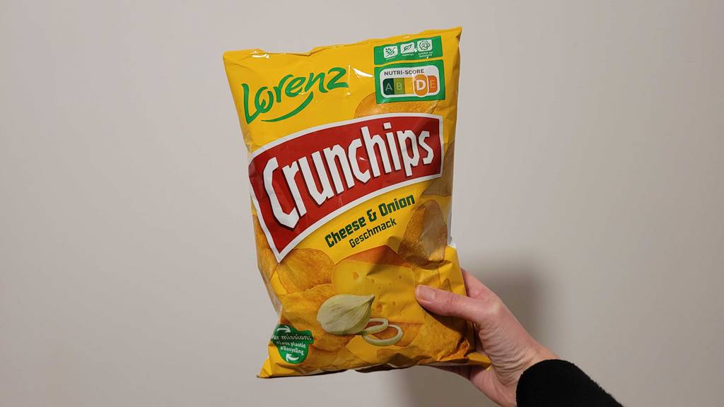 Eine Hand hält eine Packung mit Käsechips im Test von vorn vor einer weißen Wand.