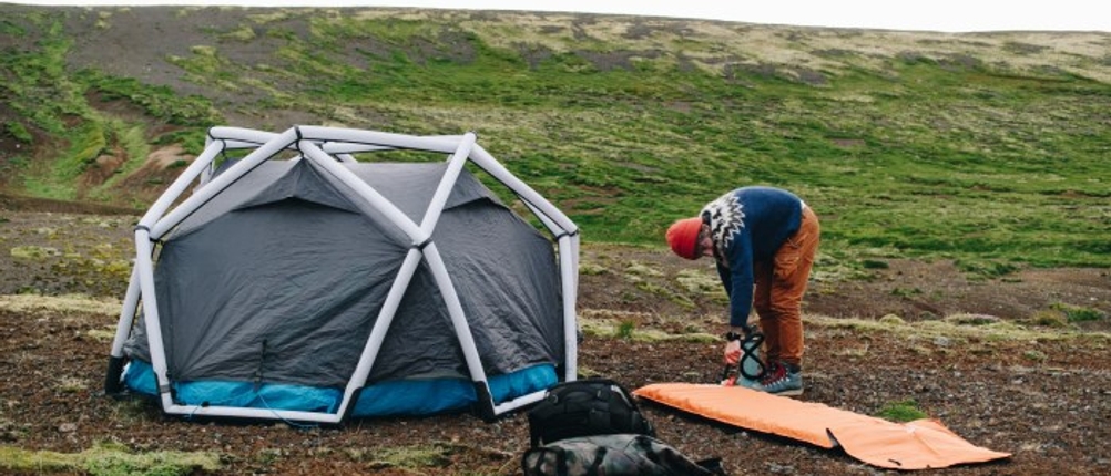 Eine Person baut eine Luftmatratze in der Natur auf, hinter ihr steht ein aufgebautes Zelt.