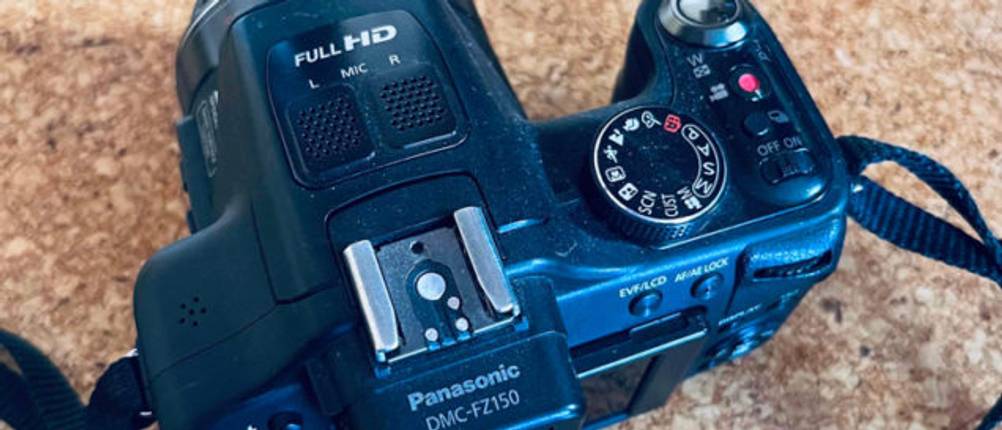 Digitalkamera-Test: Ansicht von oben einer schwarzen Full-HD-Digitalkamera mit Blick auf Einstellrad und Schultergurt auf Korkunterlage