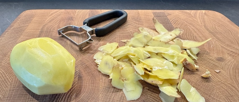 Eine geschälte Kartoffel neben dem Sparschäler und den Kartoffelschalen.