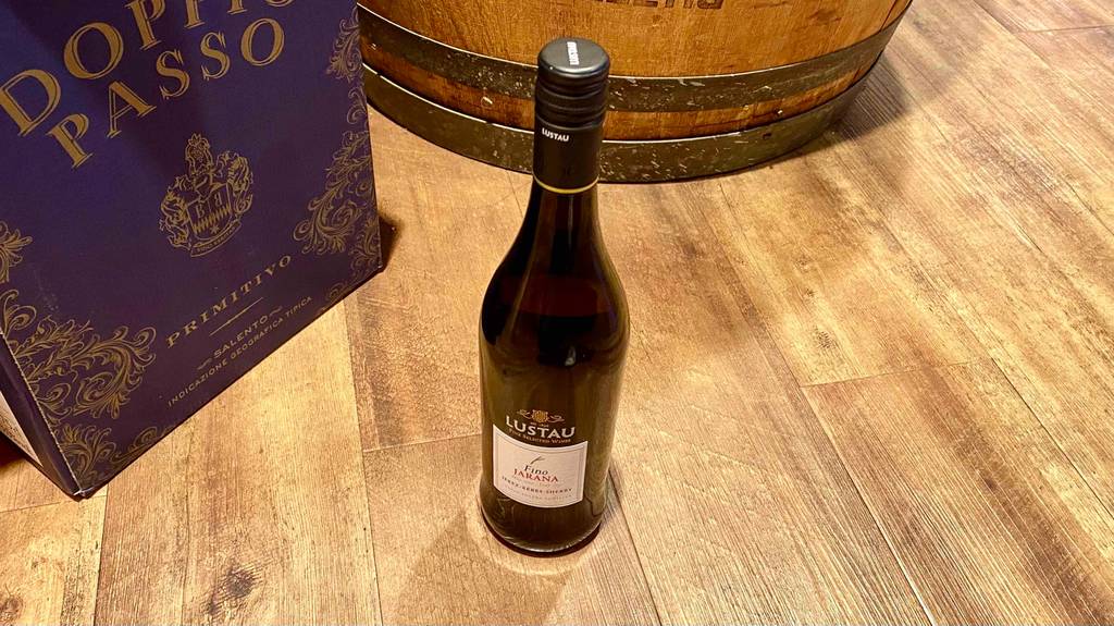Sherry-Test: eine blickdichte Flasche mit schwarzem Schraubverschluss der Marke Lustau Fino Jarana steht auf einem Holzboden vor einem Holzfass und einem Karton inklusive Primitivo Wein.