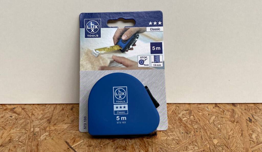 Ein getestetes Bandmaß der Marke LUX befindet sich in seiner Verpackung auf einer Pressholzplatte vor einem weißen Hintergrund.
