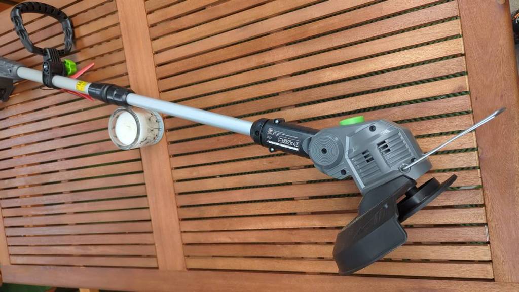 Ein getesteter Akku-Rasentrimmer liegt mit Fokus auf den Rotorkopf, den ausziehbaren Stiel sowie den flexibel einstellbaren Handgriff auf einem Holztisch neben einer weißen Kerze.