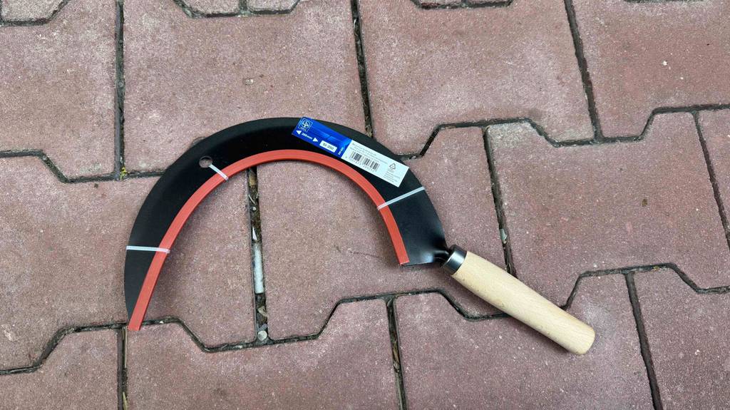Getestet: Eine runde Sichel mit Handgriff aus Holz liegt auf einem roten Steinbelag.