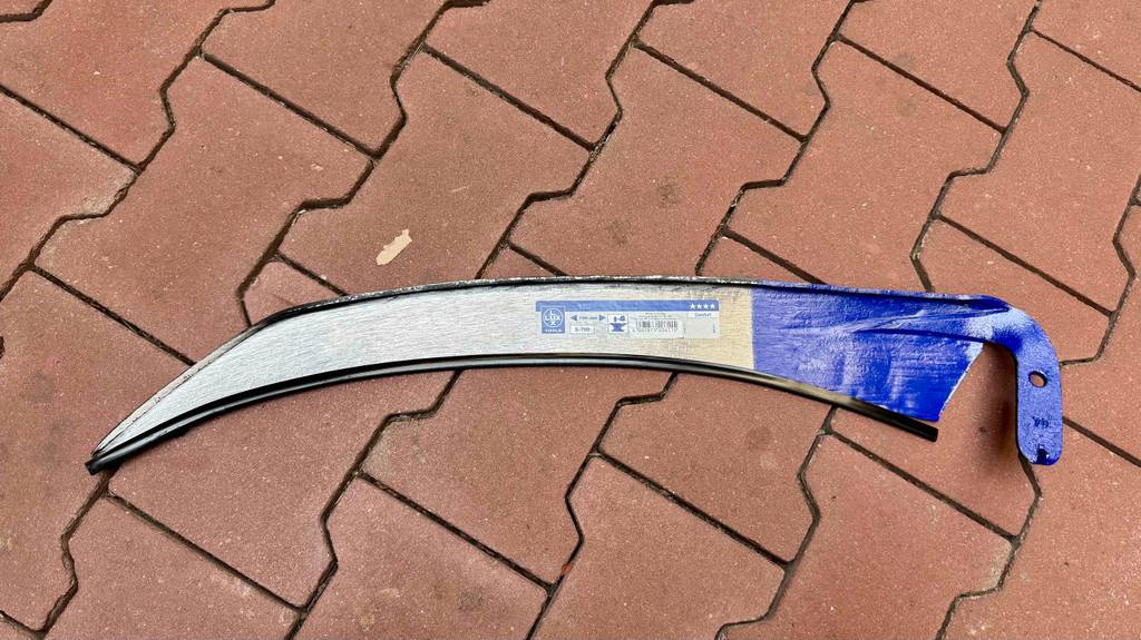 Ein getestetes Sensen-Blatt liegt inklusive Klingenschutz auf einem roten Steinbelag.