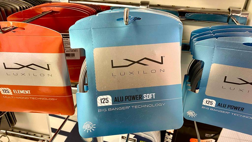 Tennissaiten im Test: blaue Packungen inklusive Tennissaiten von Luxilon der Stärke 125 Alu Power Soft hängen mit anderen Tennissaiten an einem Verkaufsstand.