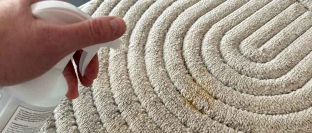 Macari-Polsterreiniger wird per Hand und Sprühkopf auf einen Fleck auf einem Teppich eingesprüht.