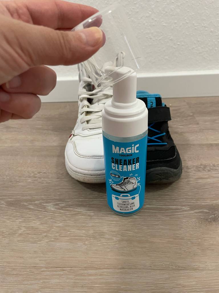 Eine Hand hält den Deckel einer Dose getesteter Sneaker-Cleaner, die vor zwei verschiedenfarbigen Schuhen steht.