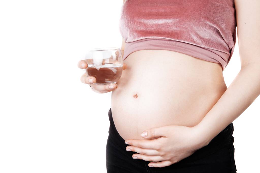 Magnesium-Ampullen-Test: Eine schwangere Frau hält ein Glas Wasser in der Hand und greift sich auf ihren Bauch. 