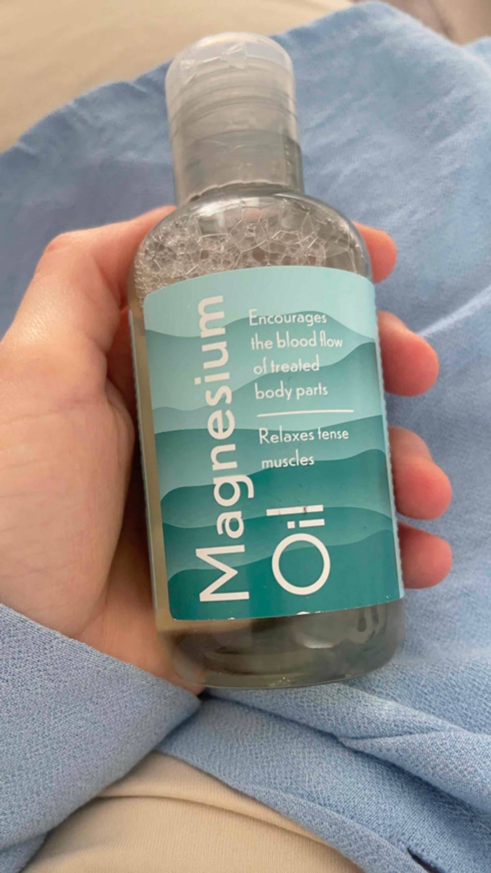 Eine Hand hält eine Flasche mit dem getesteten Magnesiumöl über einem hellblauen Gewebe.