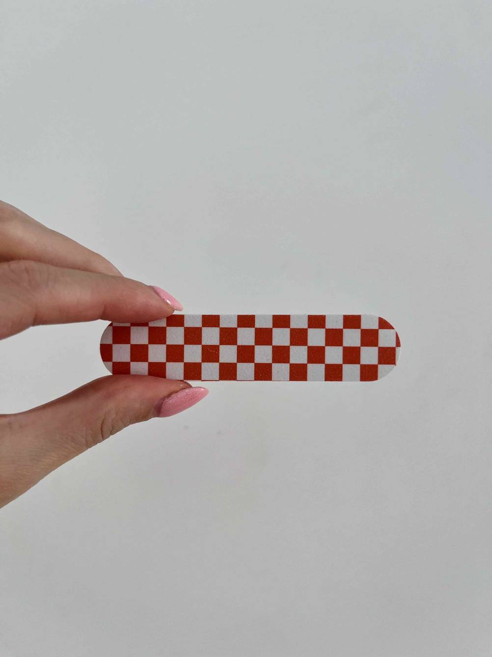 Hand hält eine rot-weiß-karierte Feile eines getesteten Nagelscheren-Sets vor einer weißen Wand.