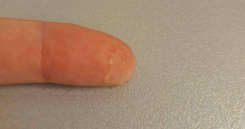 Nahaufnahme eines transparenten Make-up-Primers im Test auf einer Fingerkuppe vor silberfarbenem Hintergrund.