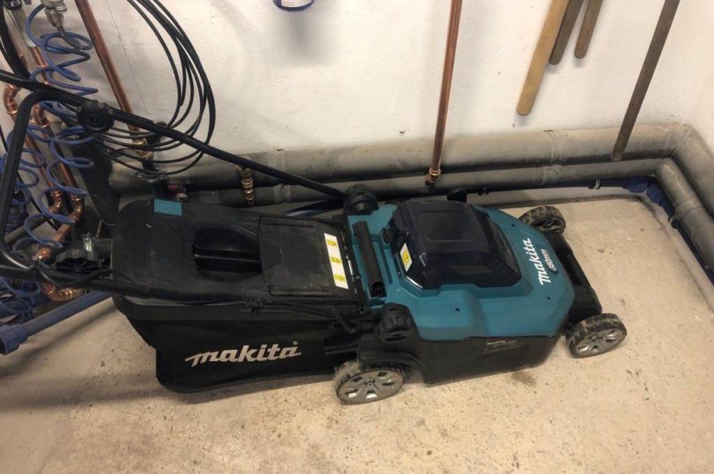 Seitenansicht von schräg oben eines getesteten Akku-Rasenmähers von Makita inklusive Auffangbeutel und höhenverstellbarem Griff auf Zementboden im Innenraum.