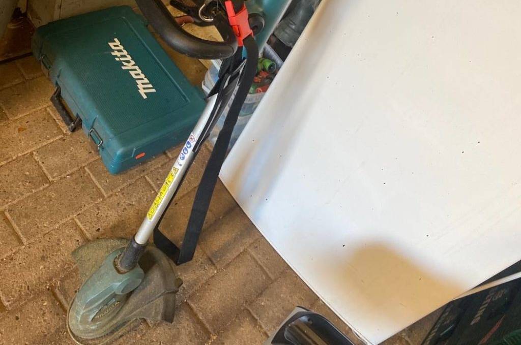 Ein Akku-Rasentrimmer im Test der Marke Makita steht zum Aufladen an einen weißen Schrank gelehnt neben einem Makita-Werkzeugkoffer in einem Handwerksraum.