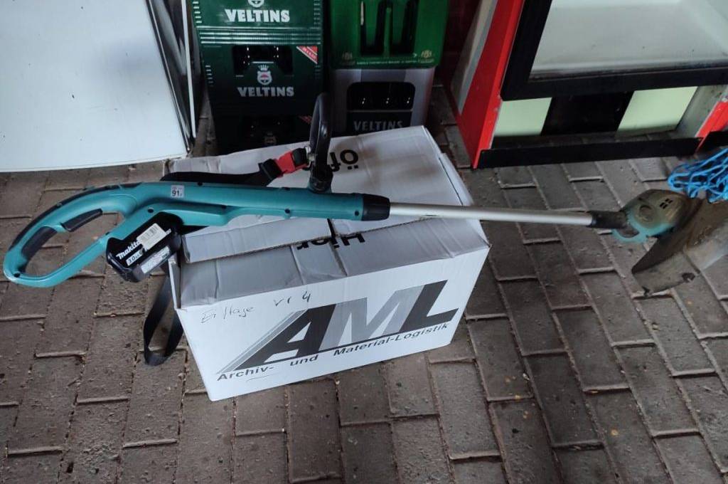 Akku-Rasentrimmer im Test der Marke Makita liegt auf einem weißen Karton in einem Garagenbereich vor Getränkekisten.