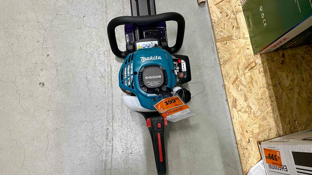Makita-Heckenscheren-Test: ein blaues, auf einem hellgrauen Zementboden liegendes Modell von oben, mit Fokus auf den Sicherheitsgriff, den Handgriff mit roten Einstellschaltern und dem Preisschild.