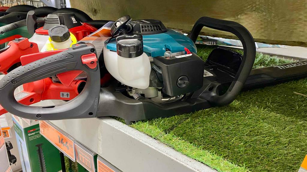 Makita-Heckenscheren im Test: ein blaues, auf einem Kunstrasen liegendes Modell im Vordergrund einer Reihe von diversen Heckenscheren. Im Fokus stehen der Tank und die Motoreinheit.