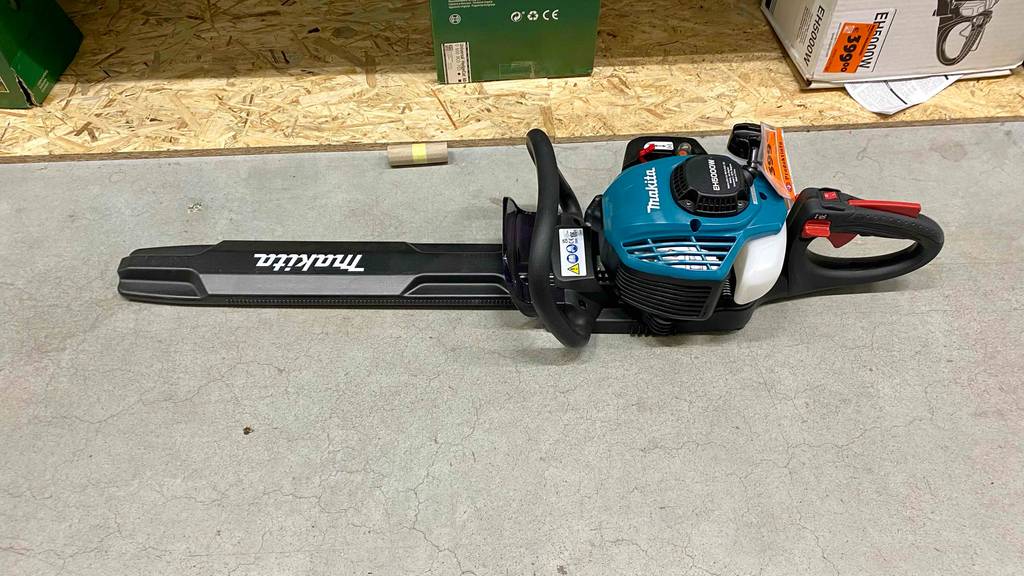 Makita-Heckenschere im Test: das blau-schwarze, auf einem hellgrauen Zementboden liegende Modell von schräg oben, mit Fokus auf das lange Sägeblatt inklusive schwarzer Abdeckung und die drei Funktionsgriffe.