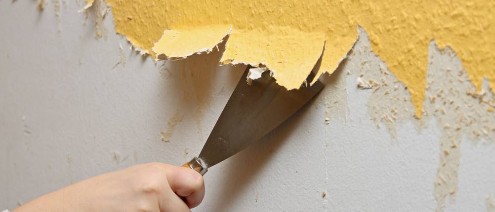 Malerset-Test: Hand mit einem Spachtel beim Entfernen gelber Tapetenreste von einer Wand
