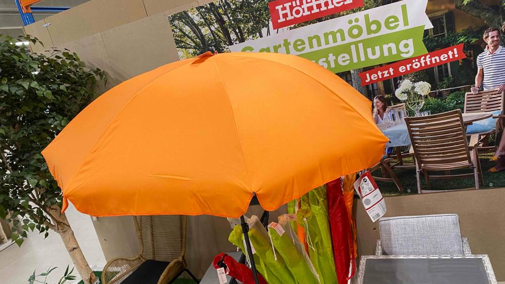 Ansicht von oben auf einen aufgespannten orangen Sonnenschirm mit Ständer im Test, der vor einem Behälter mit verpackten grünen und roten Sonnenschirmen steht.