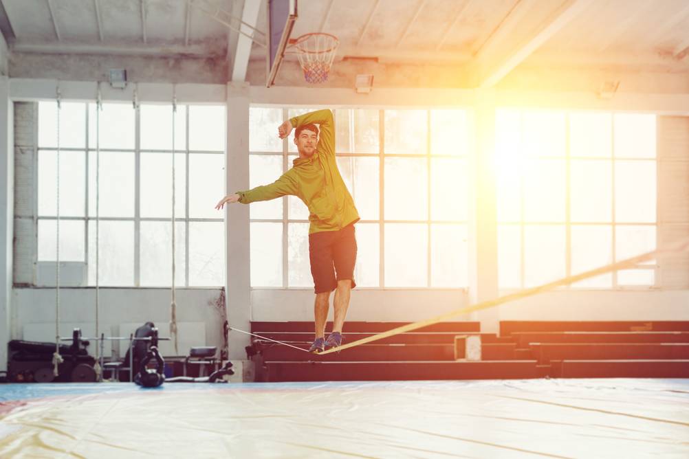 Ein Mann trainiert in einer Sporthalle auf einem getesteten Slackline-Gestell. Die Sonne leuchtet durchs Fenster. 