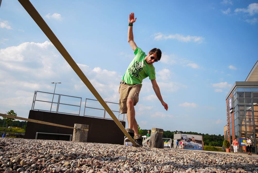 Slackline-Gestelle im Test: Ein junger Mann in grünem T-Shirt und Cargohose trainiert auf einem Parkplatz auf einer Slackline, die an Betonpfosten befestigt ist. Im Hintergrund sieht man einen Skate-Park.
