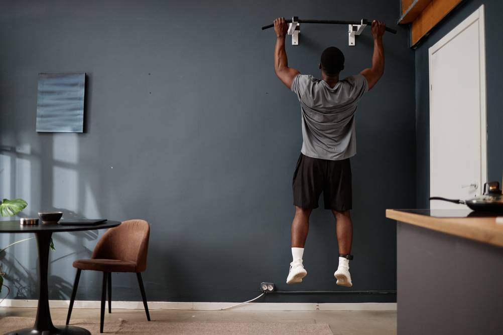 Sportgeräte für zuhause im Test: Ein Mann mit Fußfessel macht Klimmzüge an einer Stange im Wohnzimmer.
