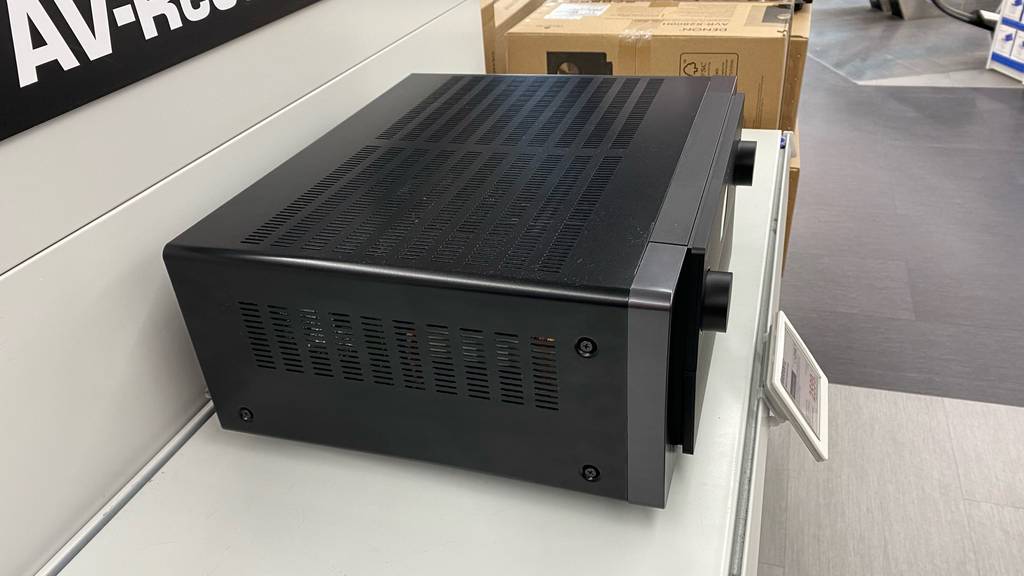 Seitenansicht von oben auf das mit Belüftungslöchern verarbeitete schwarze Gehäuse von einem AV-Receiver im Test, der auf einem Regal im Geschäft steht.