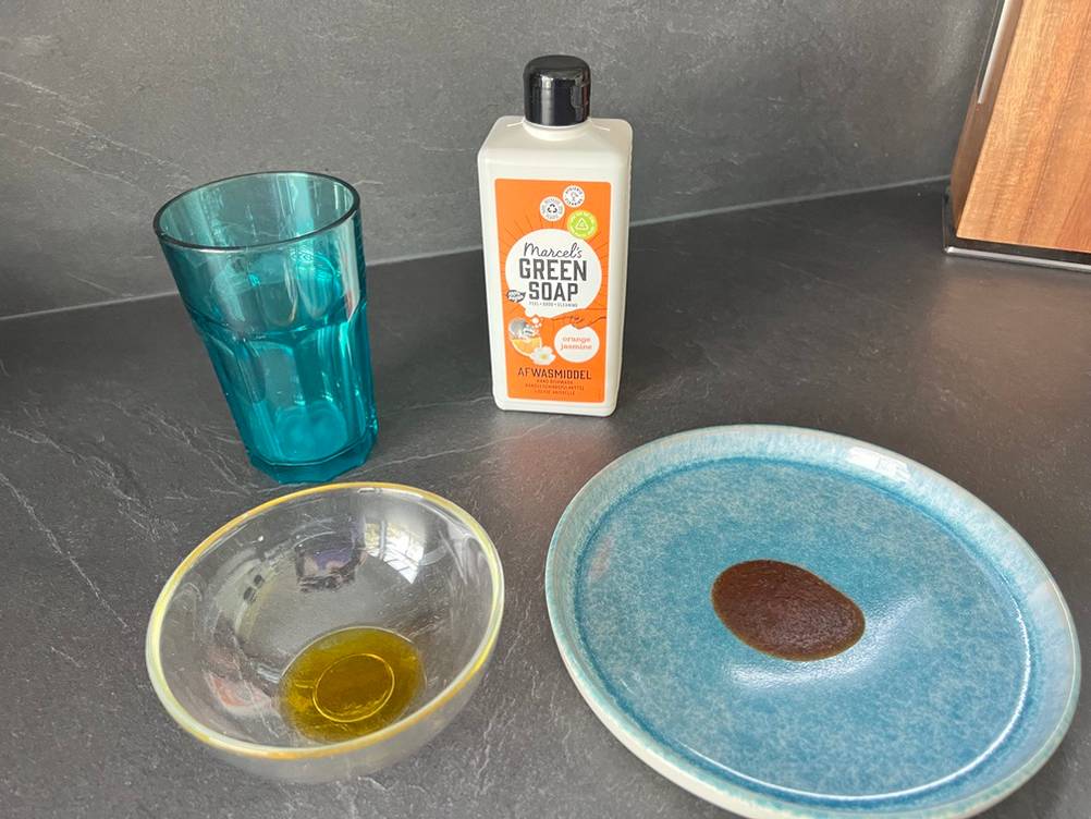 Das Marcel's Green Soap Spülmittel steht zusammen mit einem Glas eingetrockneter Milch, einer Schale Olivenöl und einem Teller mit eingetrocknetem Ketchup auf einer Küchenarbeitsplatte