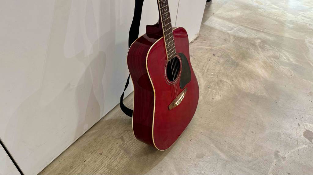 Eine bordeauxrote 7-8-Gitarre im Test lehnt an einer weißen Wand.