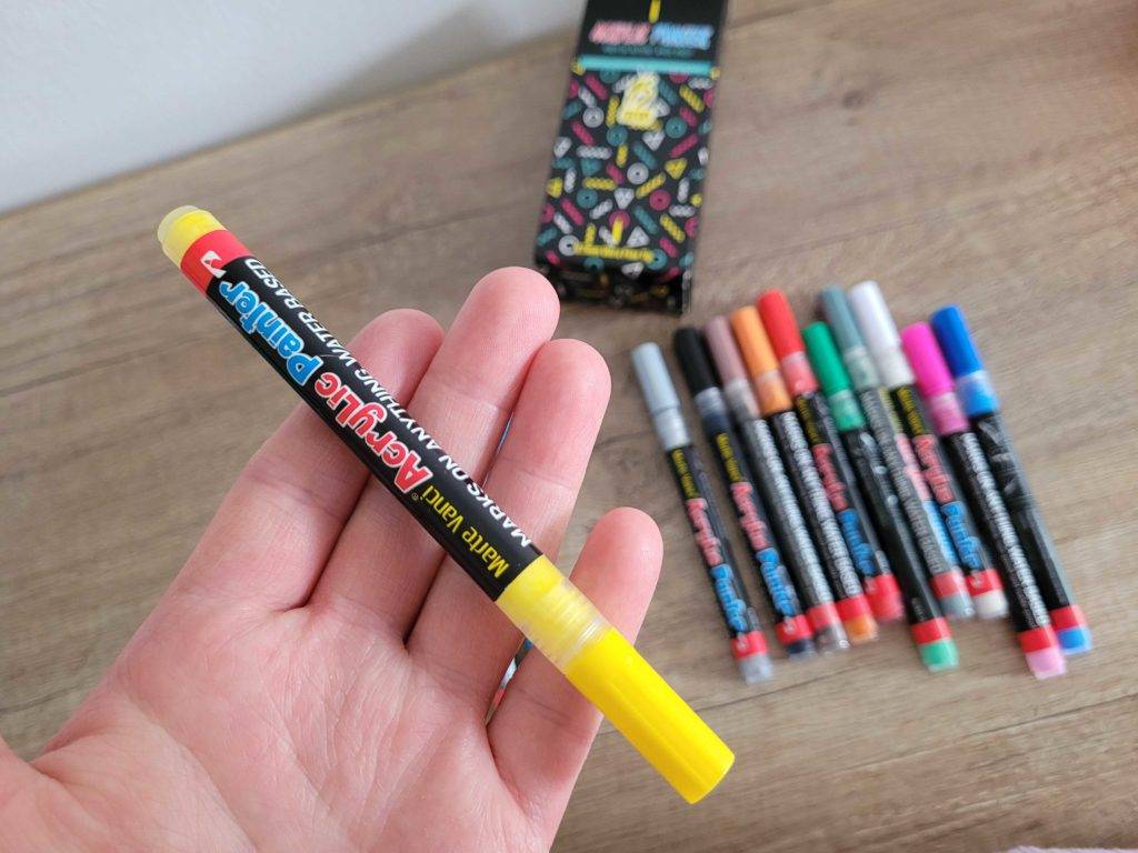 Acrylstifte getestet: Eine Hand hält einen gelben Stift über eine Holzfläche mit mehreren anderen Stiften.