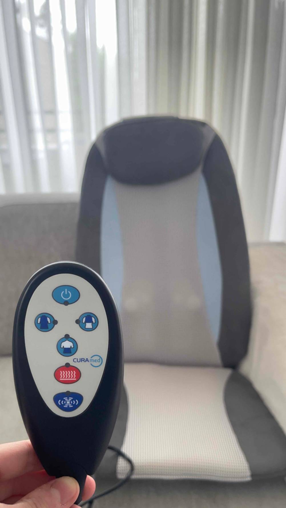 Eine Hand hält die Fernbedienung der getesteten Massagesitzauflage, im Hintergrund die Auflage.