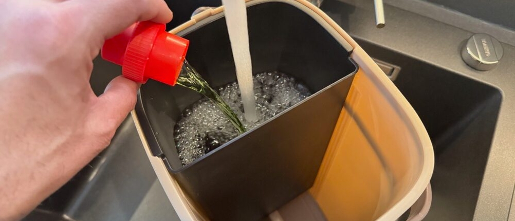 Eine Hand schütter Reiniger in den Frischwasserbehälter des Masthome-Wischmopp-Sets, während er vollläuft.