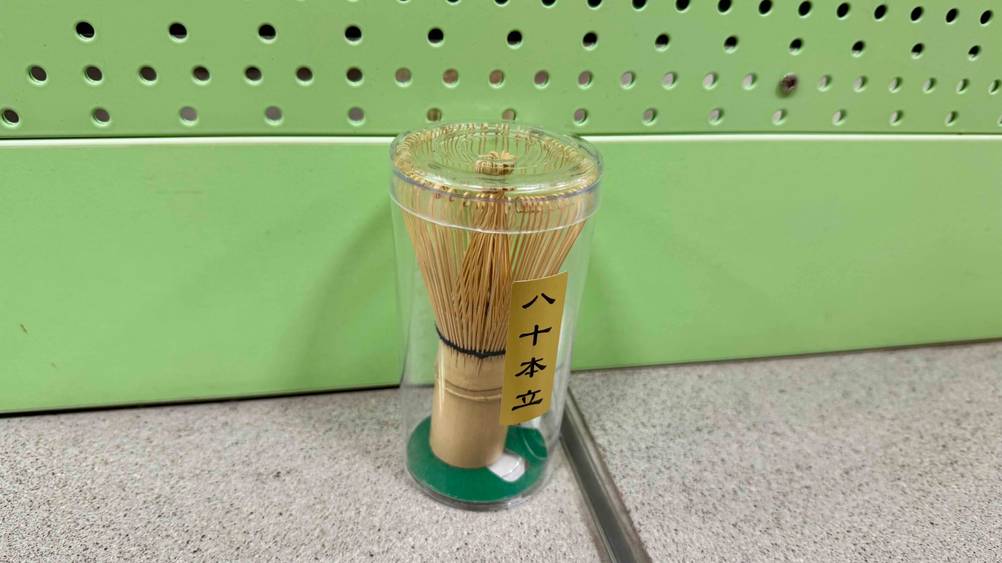 Vor einer grünen Wand steht ein verpackter Matcha-Besen im Test auf dem Boden.