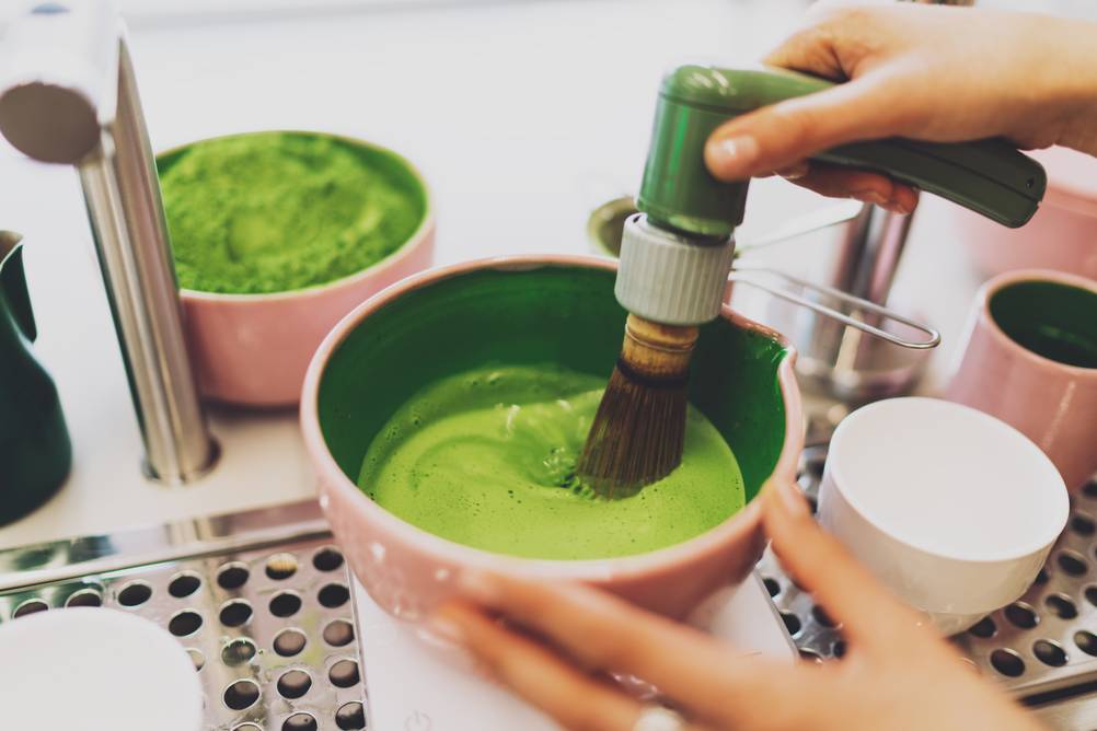 Matcha-Besen-Test: Mit einem elektrischen Matcha-Besen wird Matcha in einer kleinen Schale auf einer Metallablage zubereitet.